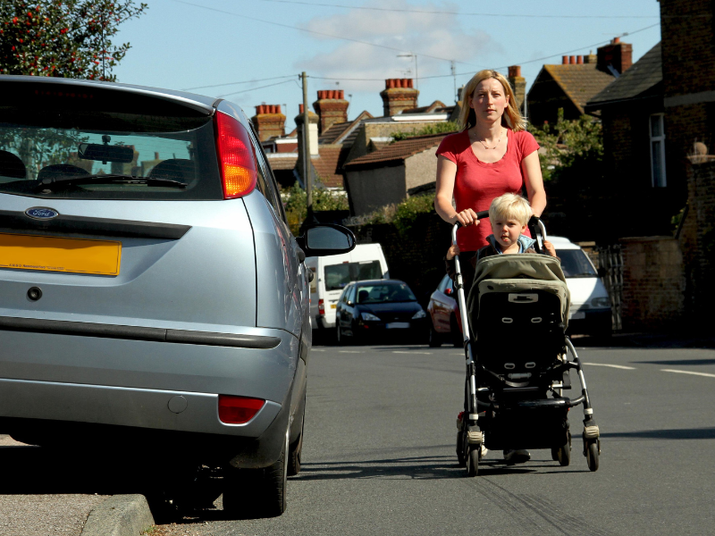 a woman walks in the road with her buggy because a car is parked on the pavement