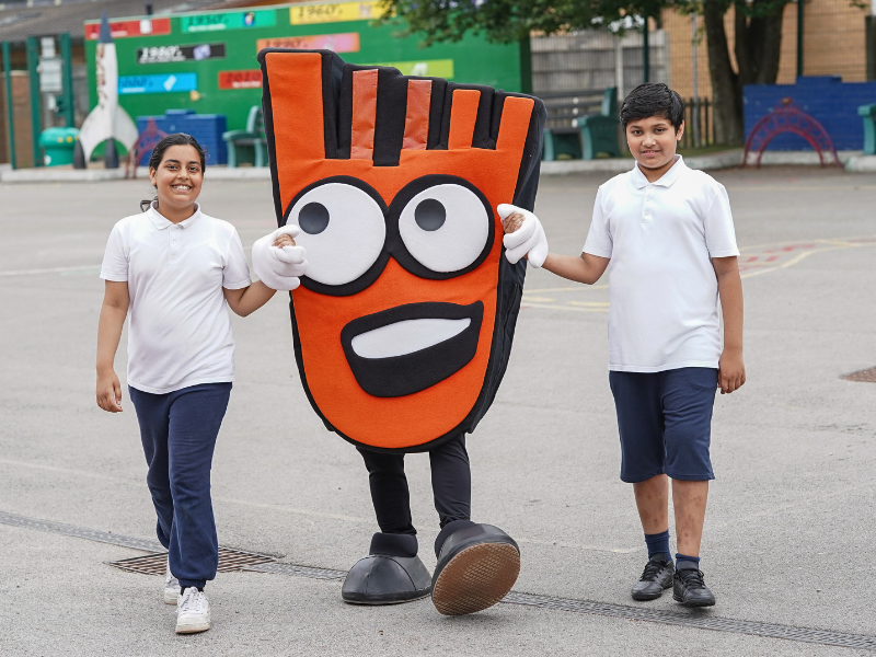two pupils hold the hands of strider the living streets mascot